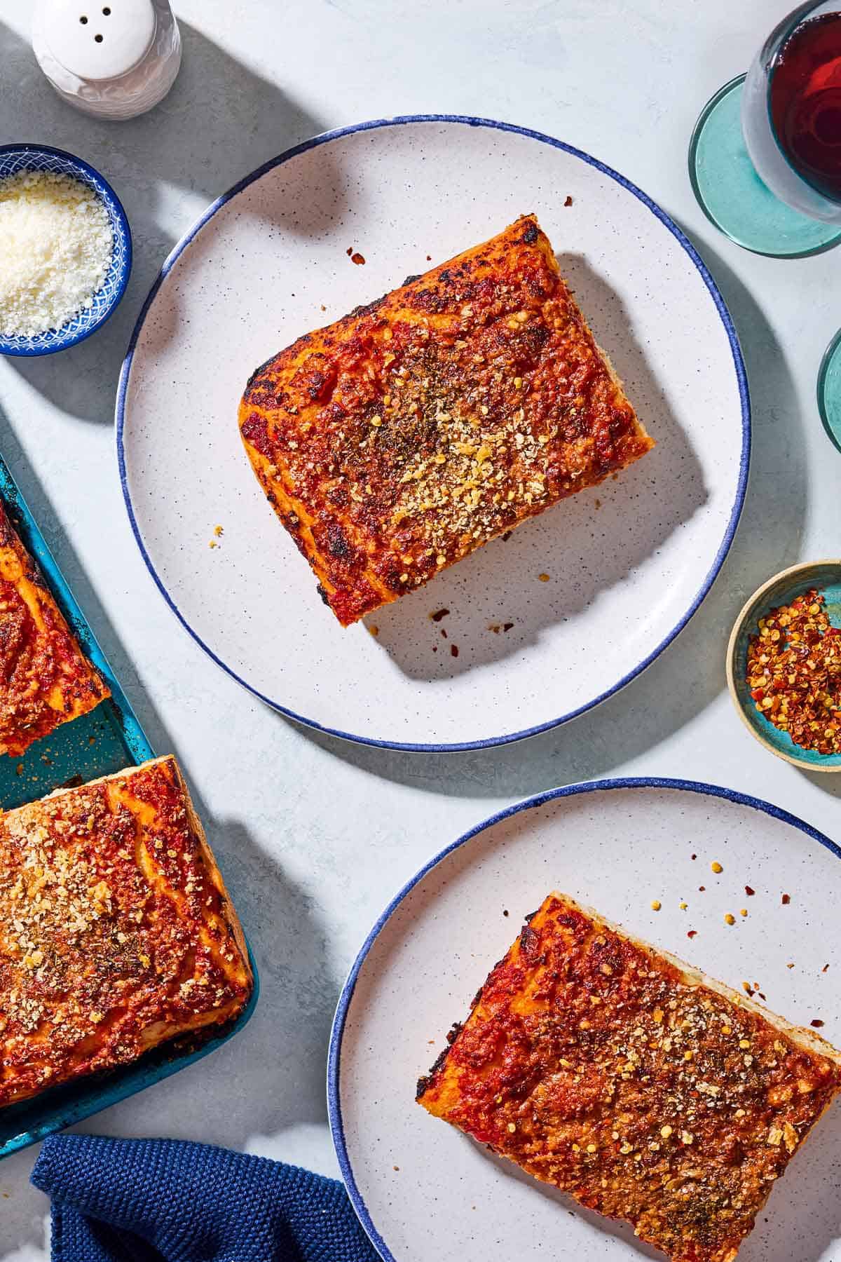 An overhead photo of 2 slices sfincione sicilian pizza on 2 plates. Next to these are small bowls pecorino cheese and red pepper flakes, a salt shaker, a cloth napkin, and a glass of red wine.