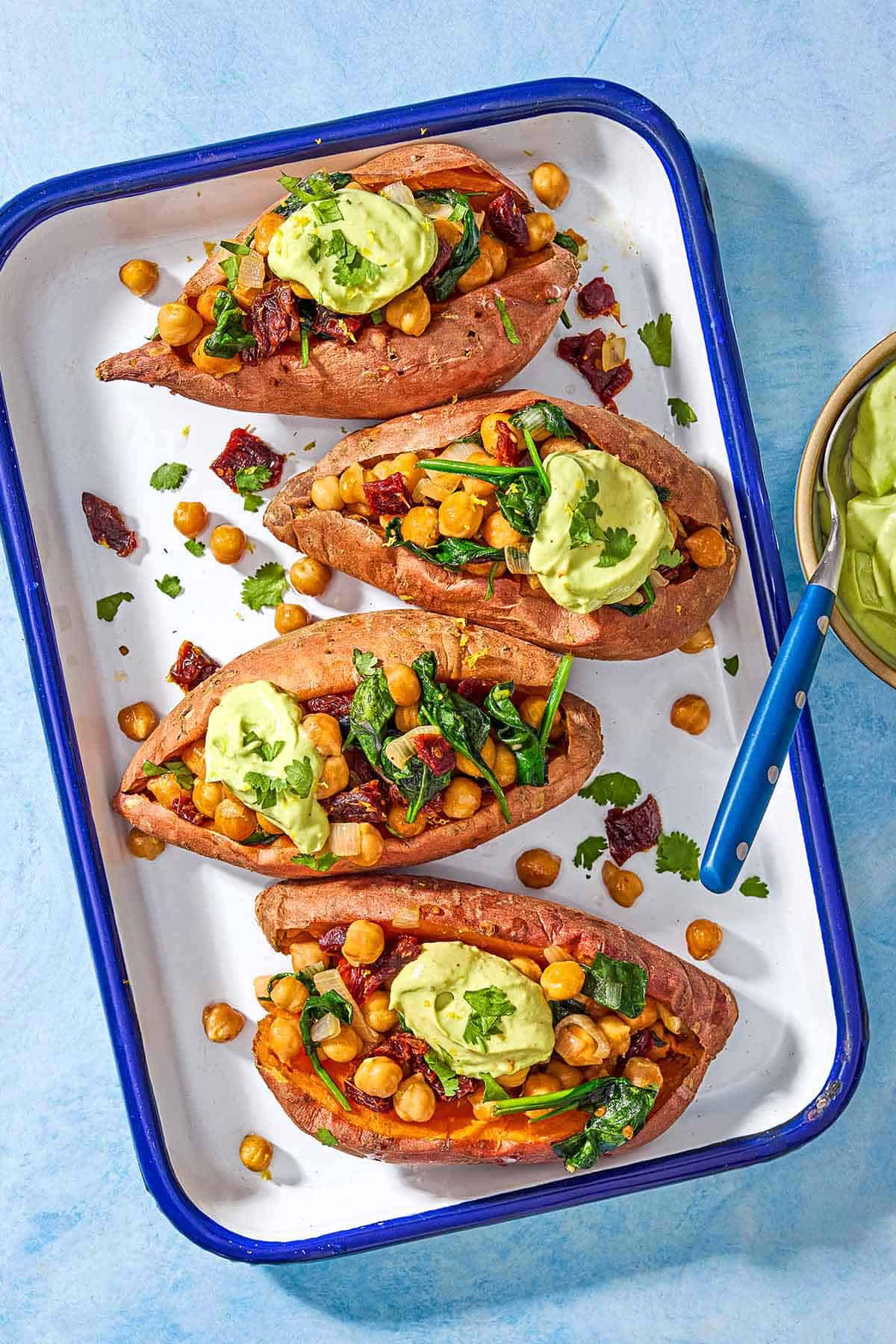 An overhead photo of 4 stuffed sweet potatoes with avocado crema on a platter next to a bowl of avocado crema with a spoon.