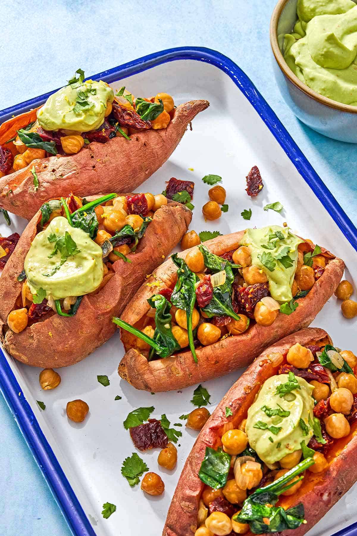 A close up of 4 stuffed sweet potatoes with avocado crema on a platter next to a bowl of avocado crema.