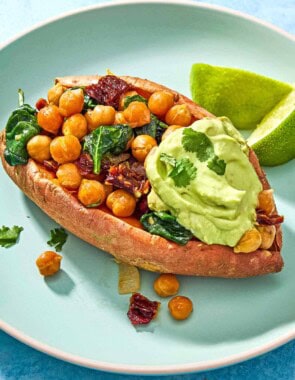 A close up of a stuffed sweet potato with a dollop of avocado crema on a plate with 2 lime wedges.