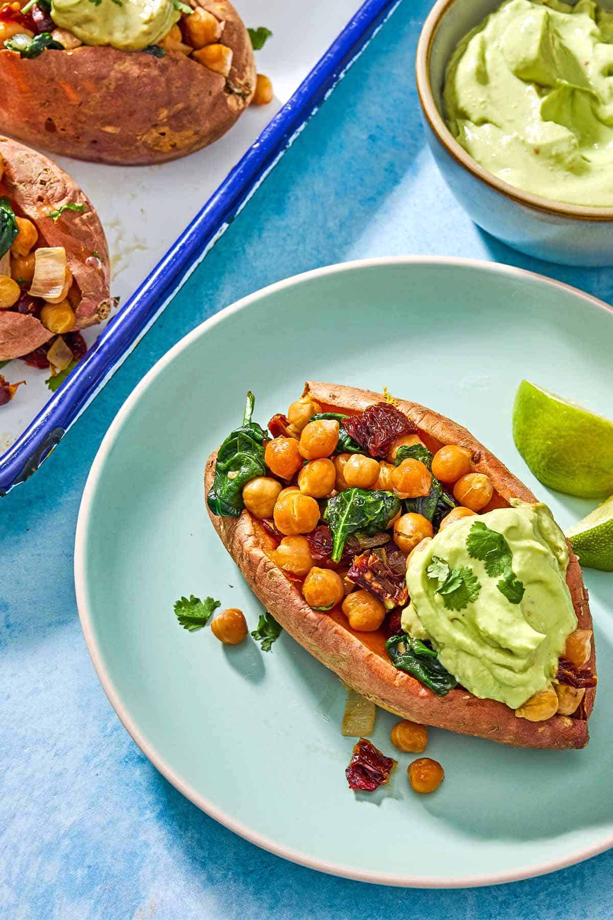 A close up of a stuffed sweet potato with a dollop of avocado crema on a plate with 2 lime wedges. Next to this is a platter with the rest of the stuffed sweet potatoes and a bowl of avocado crema.