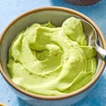 A close up of avocado crema in a bowl with a spoon. Next to this is a half of an avocado and a small bowl of aleppo pepper.