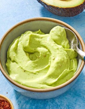 A close up of avocado crema in a bowl with a spoon. Next to this is a half of an avocado and a small bowl of aleppo pepper.