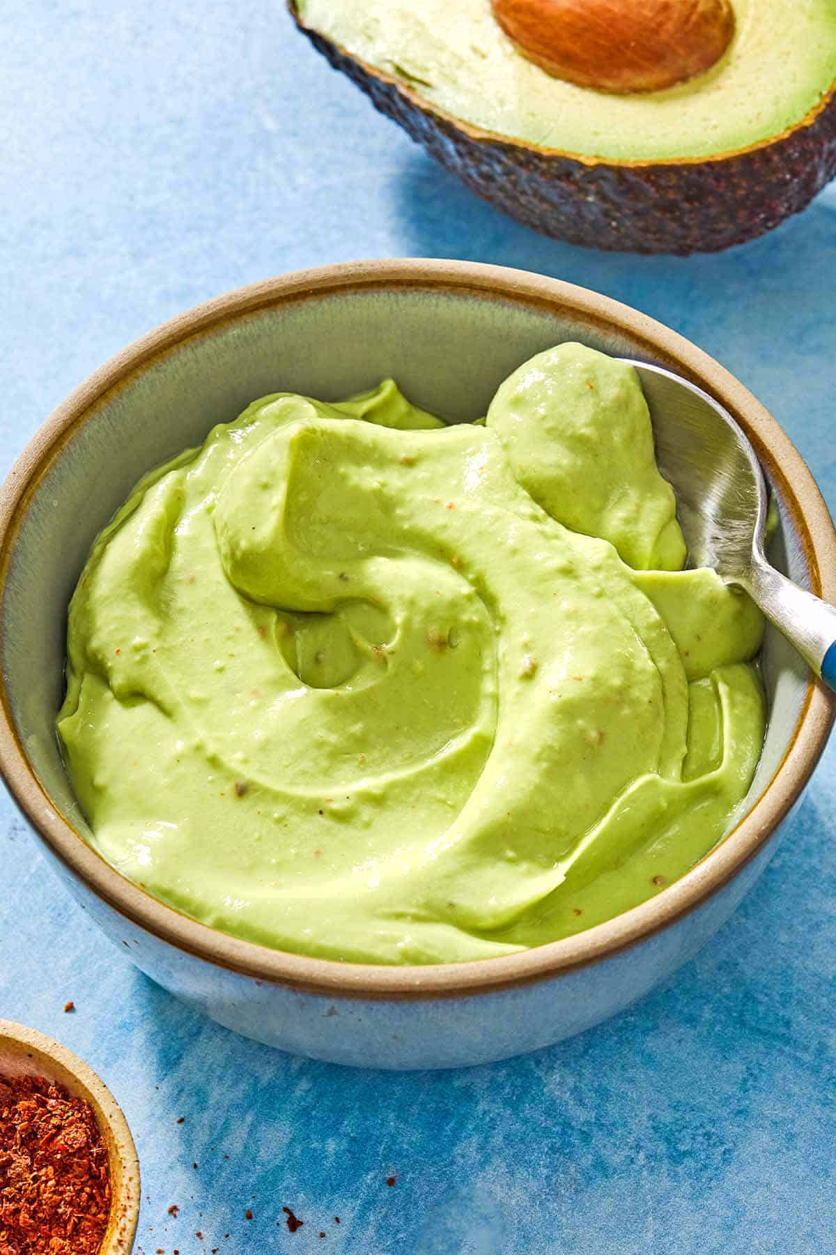 A close up of avocado crema in a bowl with a spoon. Next to this is a half of an avocado and a small bowl of aleppo pepper.