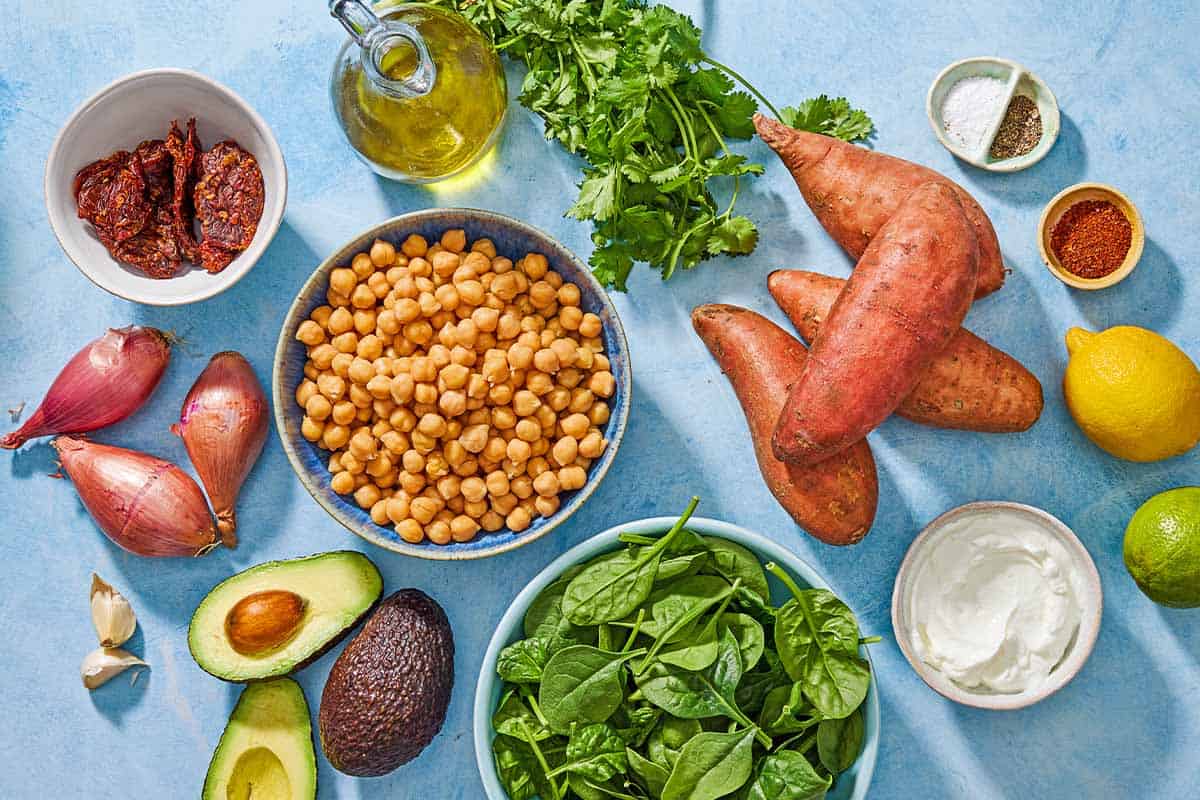 Ingredients for stuffed sweet potatoes with avocado crema including sweet potatoes, olive oil, chickpeas, shallots, garlic, sundried tomatoes, baby spinach, lime, salt, black pepper, cilantro, avocados, greek yogurt, lemon, lime, and aleppo pepper.