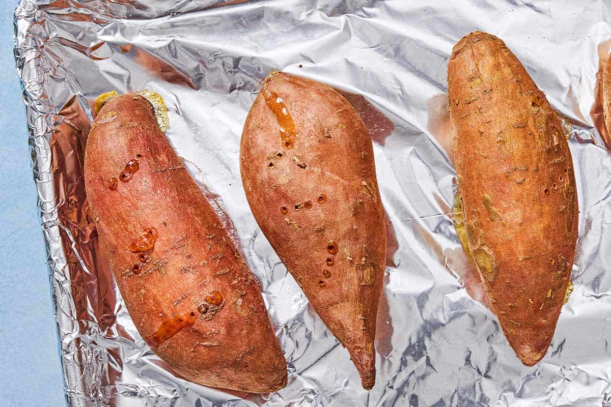 A close up of 3 sweet potatoes on a foil-lined sheet pan.