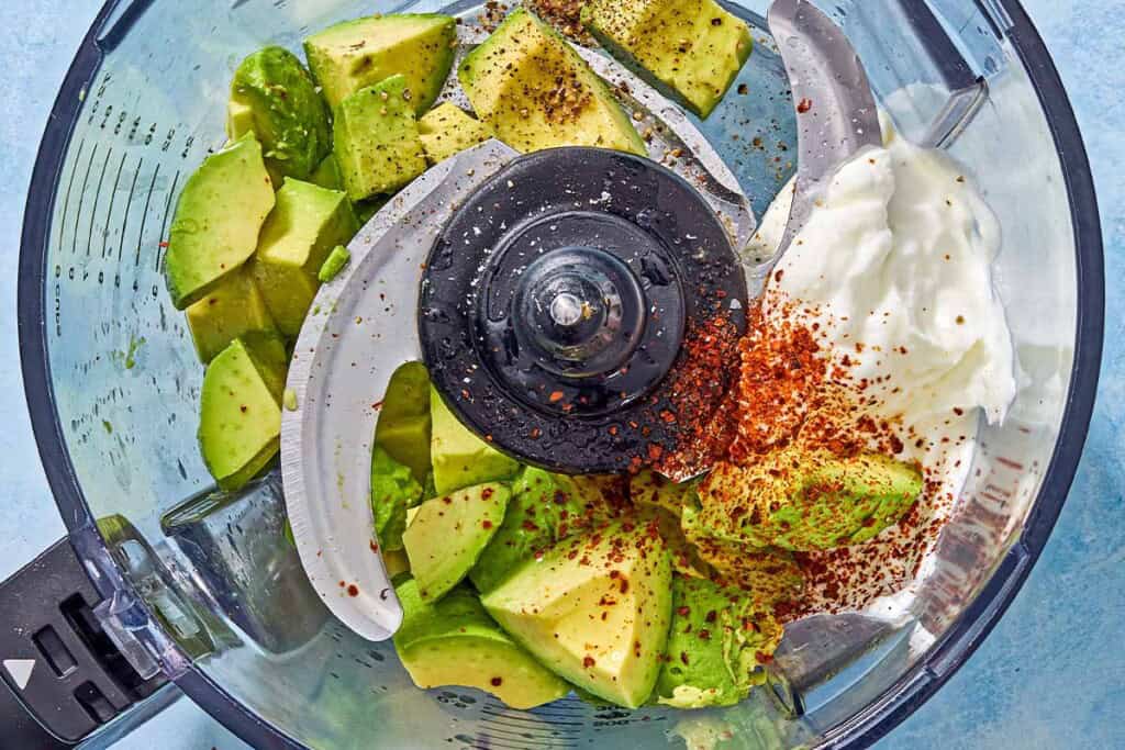 An overhead photo of the ingredients for the avocado crema in the bowl of a food processor fitted with a blade just before being mixed together.