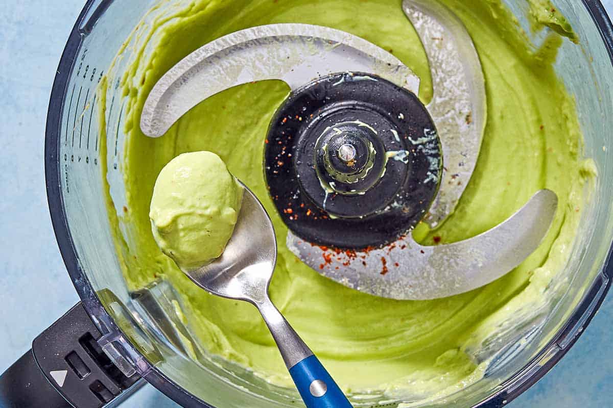 A close up photo of a avocado crema on a spoon being held above the rest of the avocado crema in the bowl of a food processor fitted with a blade.