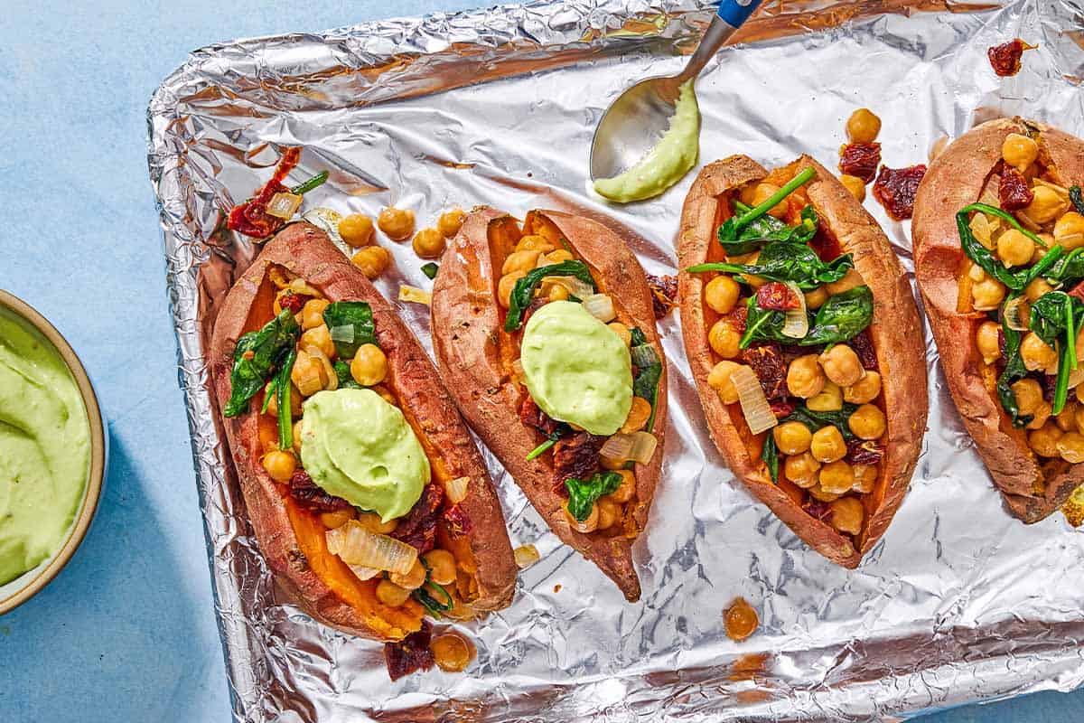 An overhead photo of 4 stuffed sweet potatoes, 2 with a dollop of avocado crema, on a foil-line sheet pan with a spoon. Next to this is a bowl of avocado crema.