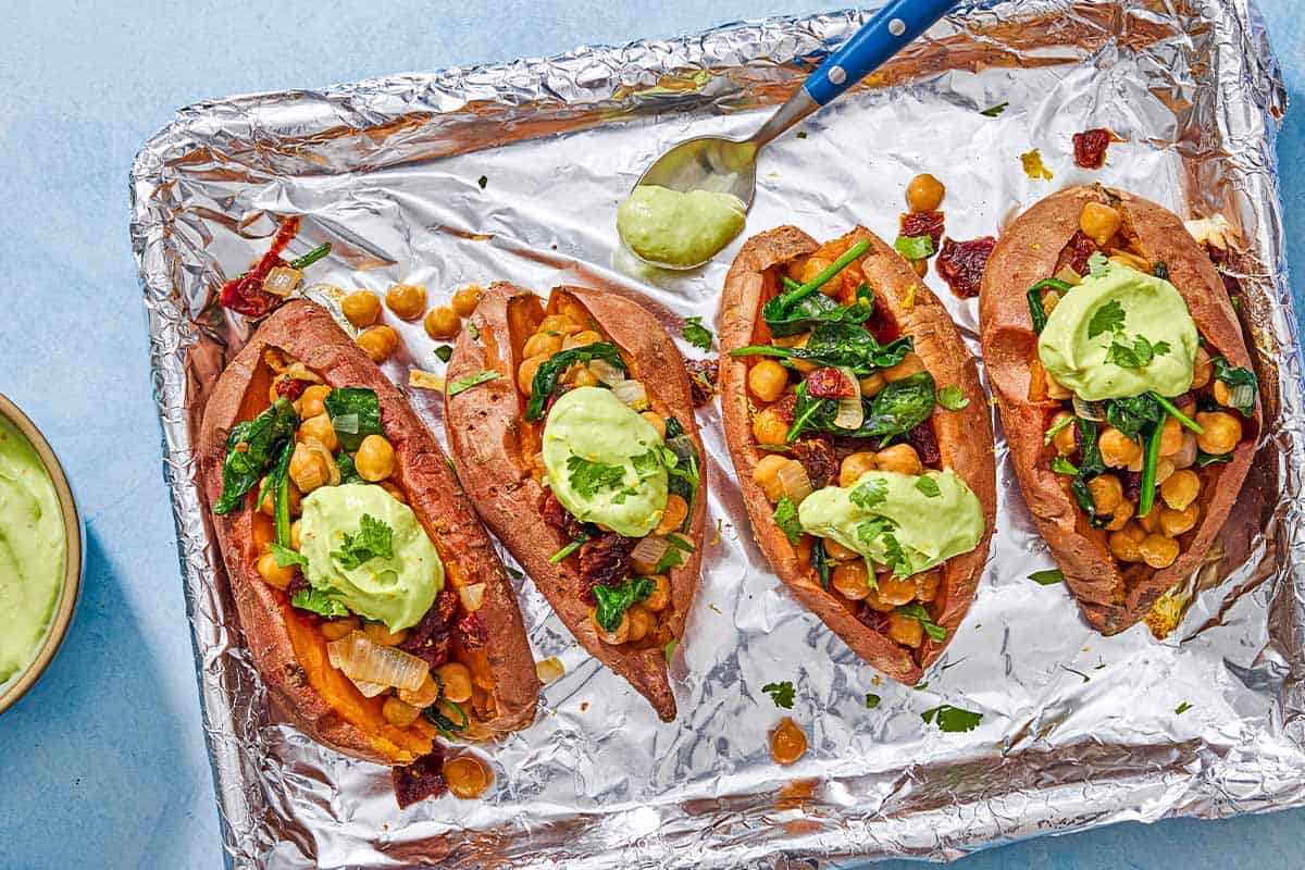 An overhead photo of 4 stuffed sweet potatoes, 2 with a dollop of avocado crema, on a foil-line sheet pan with a spoon. Next to this is a bowl of avocado crema.