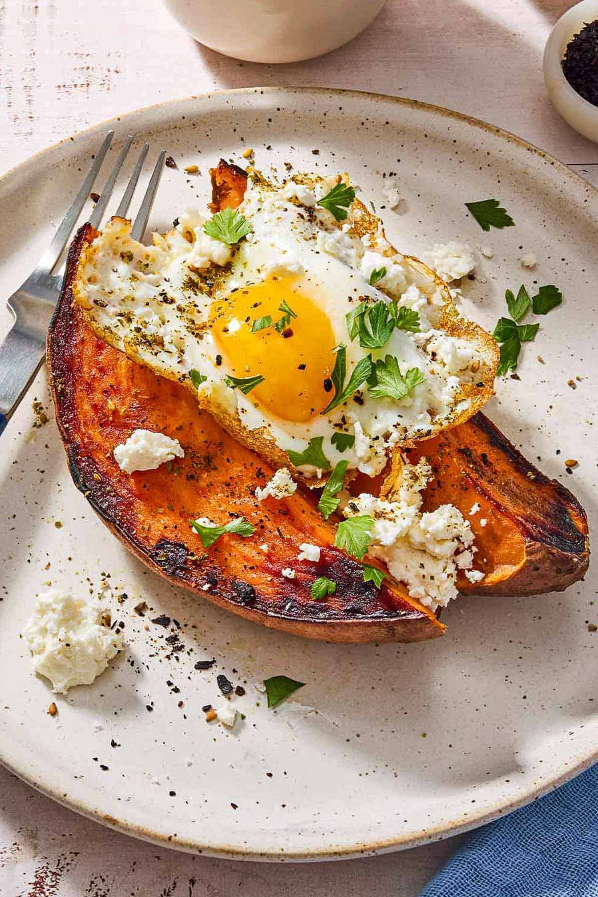 A close up of a sweet potato topped with a fried egg, spices, feta and parsley on a plate with a fork.