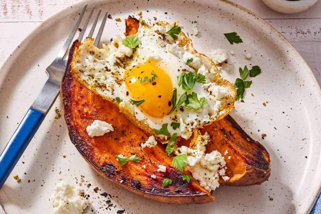 A close up of a sweet potato topped with a fried egg, spices, feta and parsley on a plate with a fork.