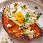 A close up of a sweet potato topped with a fried egg, spices, feta and parsley on a plate with a fork.