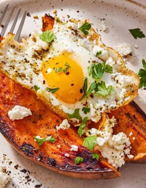 A close up of a sweet potato topped with a fried egg, spices, feta and parsley on a plate with a fork.