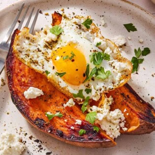 A close up of a sweet potato topped with a fried egg, spices, feta and parsley on a plate with a fork.