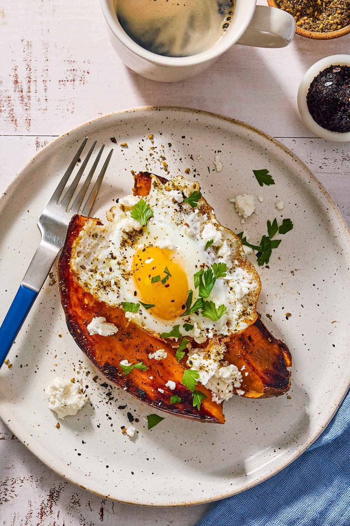 An overhead photo of a sweet potato topped with a fried egg, spices, feta and parsley on a plate with a fork. Next to this is a cup of coffee, a cloth napkin, a cup of coffee, and bowls of za'atar and urfa biber.