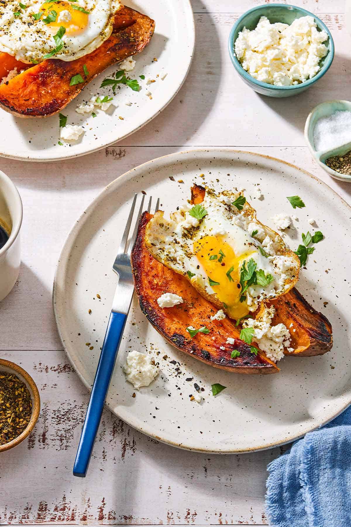 A close up of a sweet potato topped with a fried egg, spices, feta and parsley on a plate with a fork. Next to this is a cloth napkin, bowls of za'atar, feta, salt and pepper, and another sweet potato topped with an egg on a plate.