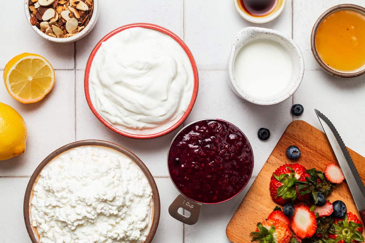 Ingredients for a yogurt parfait recipe including sliced almonds, cottage cheese, honey, lemon, vanilla extract, greek yogurt, milk, berry compote and fresh berries.