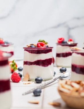 A close up of a yogurt parfait in a glass jar topped with berries. Next to this is a spoon, several more jars of the parfait, a bowl of almonds, and various berries scattered about.