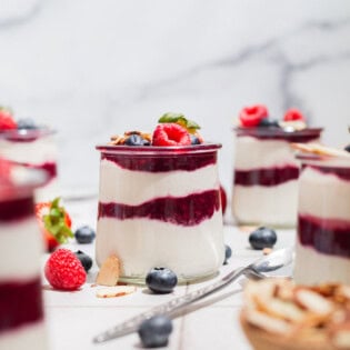 A close up of a yogurt parfait in a glass jar topped with berries. Next to this is a spoon, several more jars of the parfait, a bowl of almonds, and various berries scattered about.