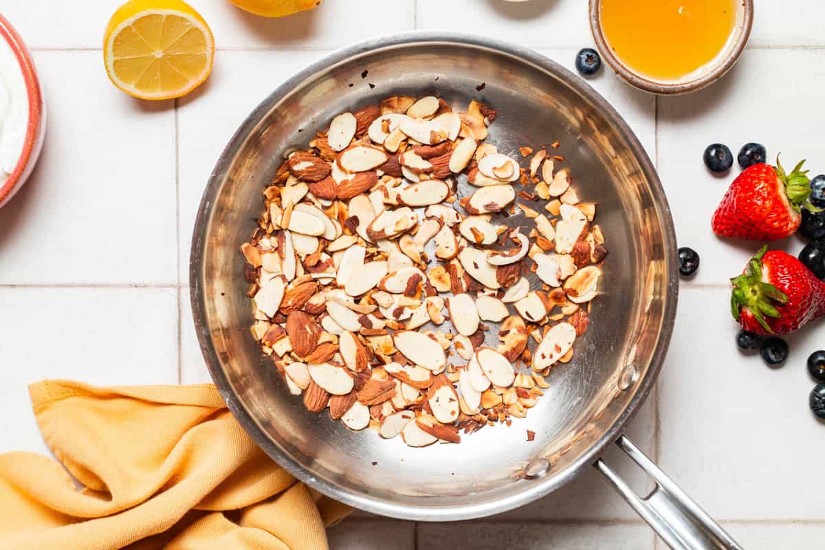 An overhead photo of sliced almonds being toasted in a skillet. Next to this is a cloth napkin, some strawberries and blueberries, a bowl of lemon juice and a lemon half.