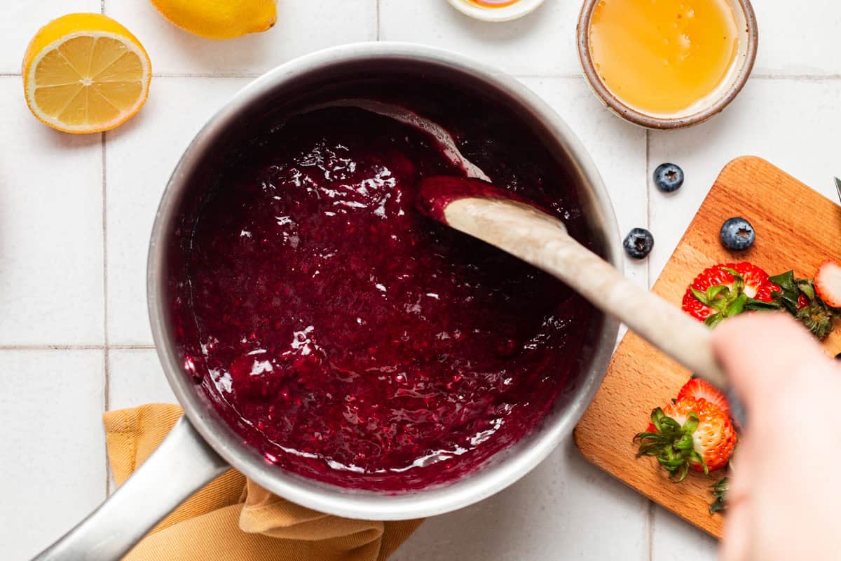 An overhead photo of berry compote in a saucepan with a wooden spoon. Next to this is a cutting board with various berries, a small bowl of lemon juice and some lemons.