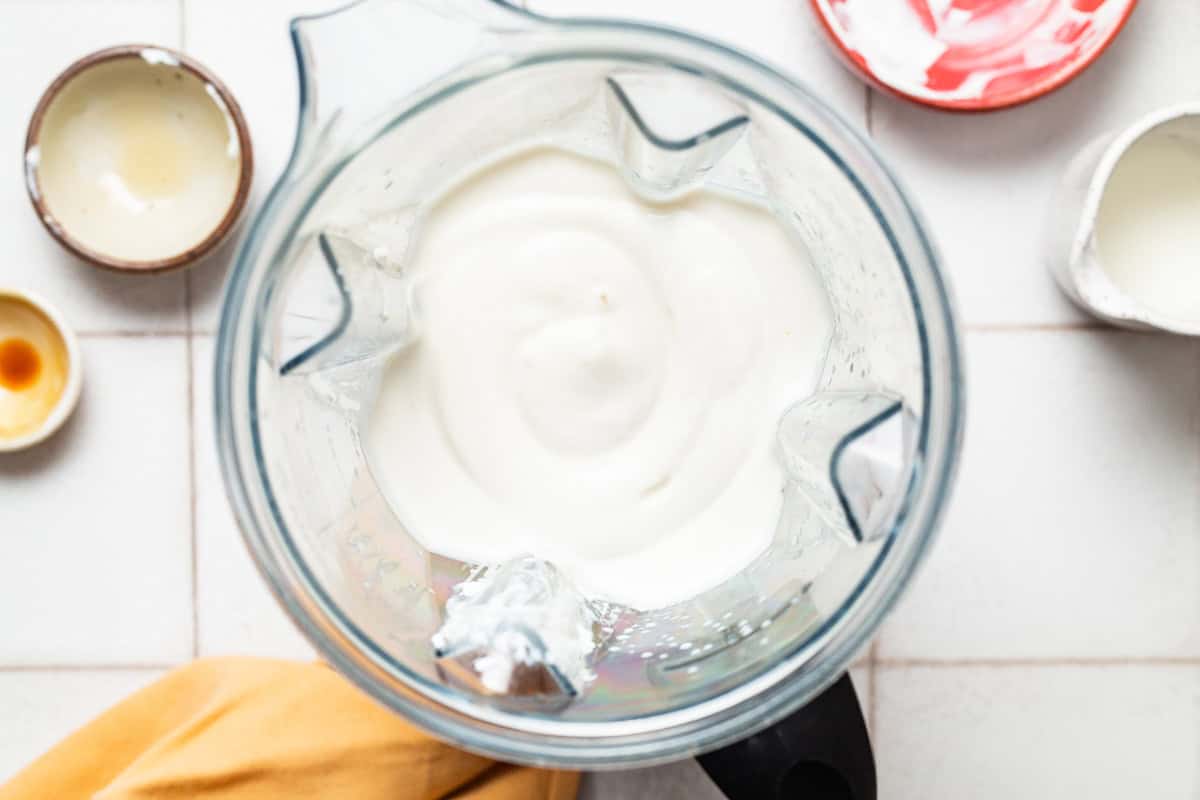 An overhead photo of the cottage cheese, honey, lemon juice, lemon zest and vanilla extract combined in a blender. Next to this are 3 empty bowls and a small pitch with a bit of milk.