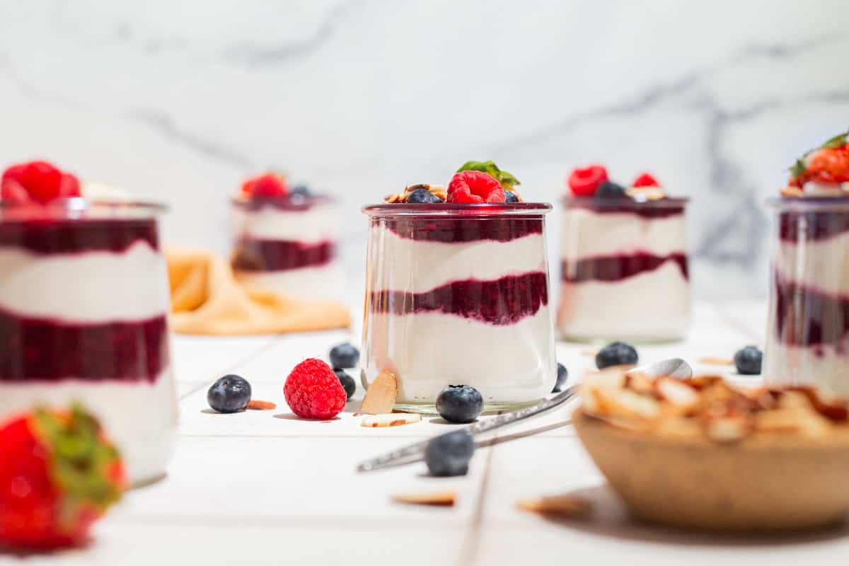 A close up of a yogurt parfait in a glass jar topped with berries. Next to this is a spoon, several more jars of the parfait, and various berries scattered about.