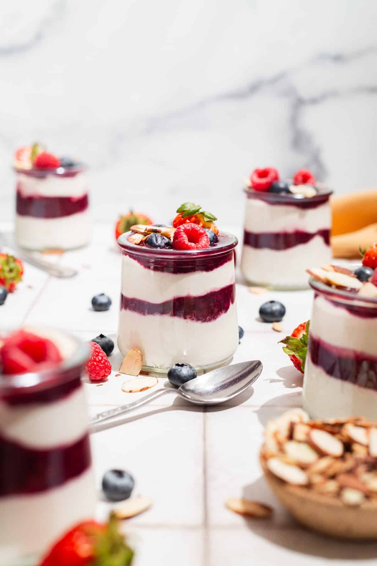 A close up of a yogurt parfait in a glass jar topped with berries. Next to this is a spoon, several more jars of the parfait, a bowl of almonds, and various berries scattered about.