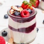 A close up of a yogurt parfait in a glass jar topped with berries. Next to this is a spoon and various berries scattered about.