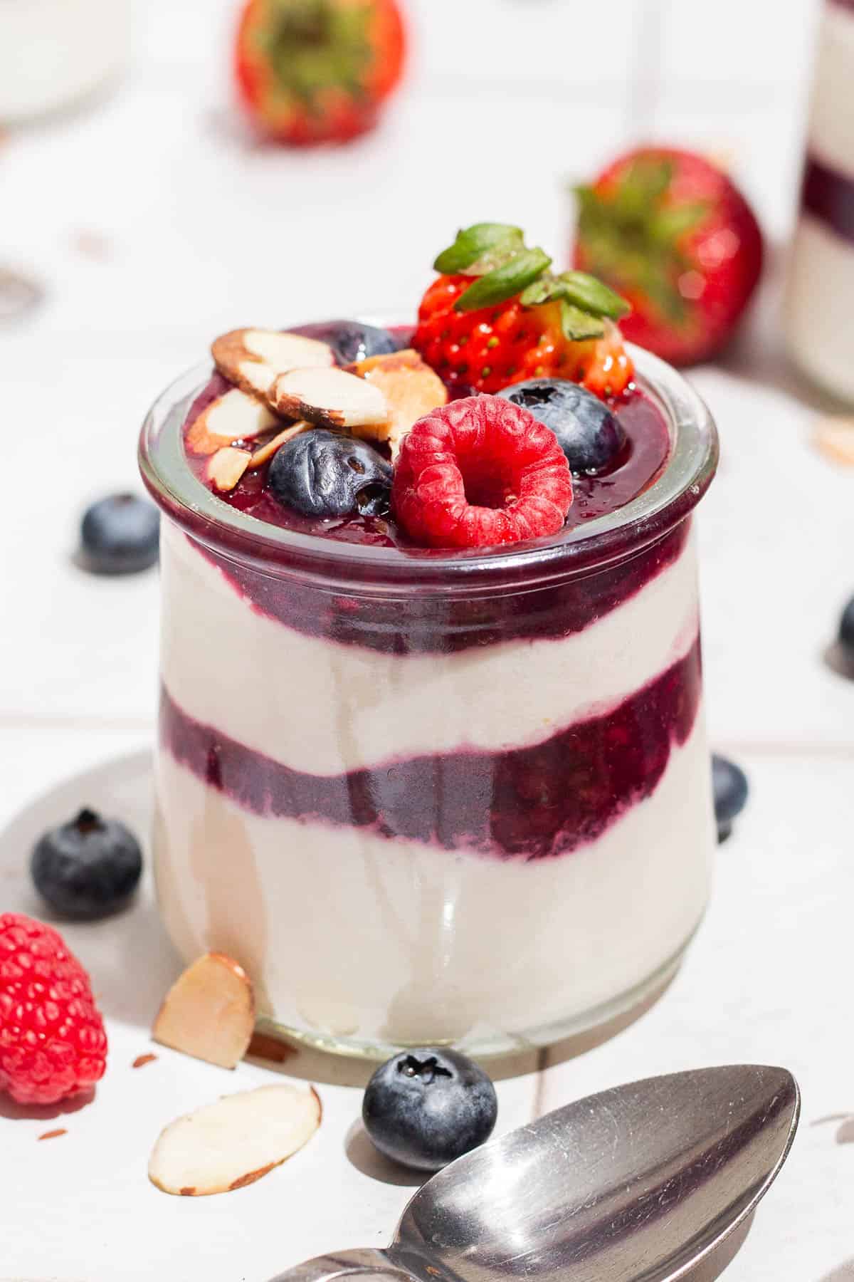 A close up of a yogurt parfait in a glass jar topped with berries. Next to this is a spoon and various berries scattered about.