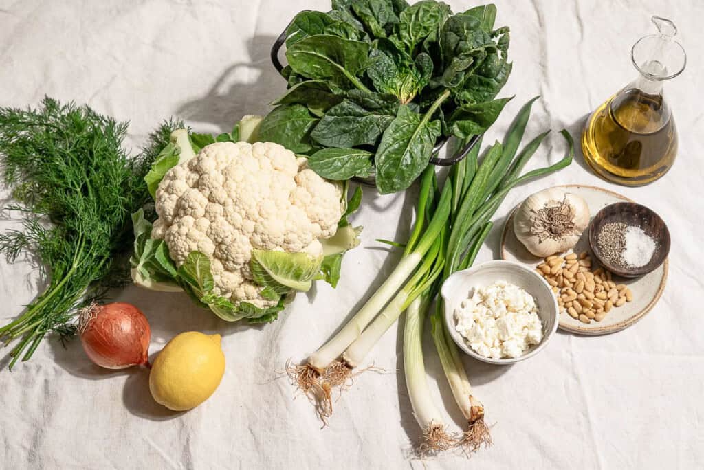 Ingredients for a spinach soup recipe including cauliflower, olive oil, salt, black pepper, garlic, onion, scallions, dill, baby spinach, lemon, feta, and pine nuts.