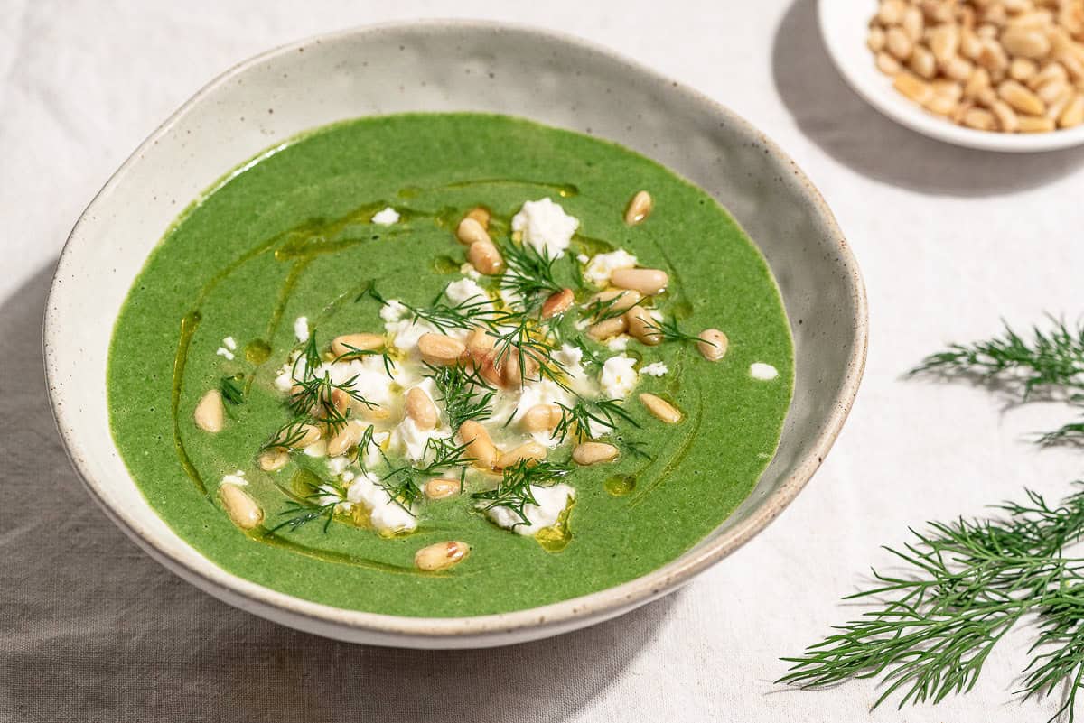 A close up of a serving of spinach soup in a bowl topped with dill, pine nuts, crumbled feta and a drizzle of olive oil. Next to this is a sprig of dill and a small bowl of pine nuts.