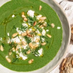 An overhead photo of a serving of spinach soup in a bowl topped with dill, pine nuts, crumbled feta and a drizzle of olive oil. Next to this is a cloth napkin and a piece of crusty bread.