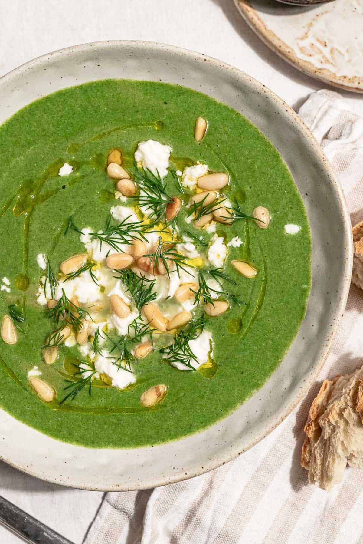 An overhead photo of a serving of spinach soup in a bowl topped with dill, pine nuts, crumbled feta and a drizzle of olive oil. Next to this is a cloth napkin and a piece of crusty bread.