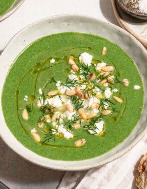 A close up of a serving of spinach soup in a bowl topped with dill, pine nuts, crumbled feta and a drizzle of olive oil. Next to this is a bowl of salt and pepper.