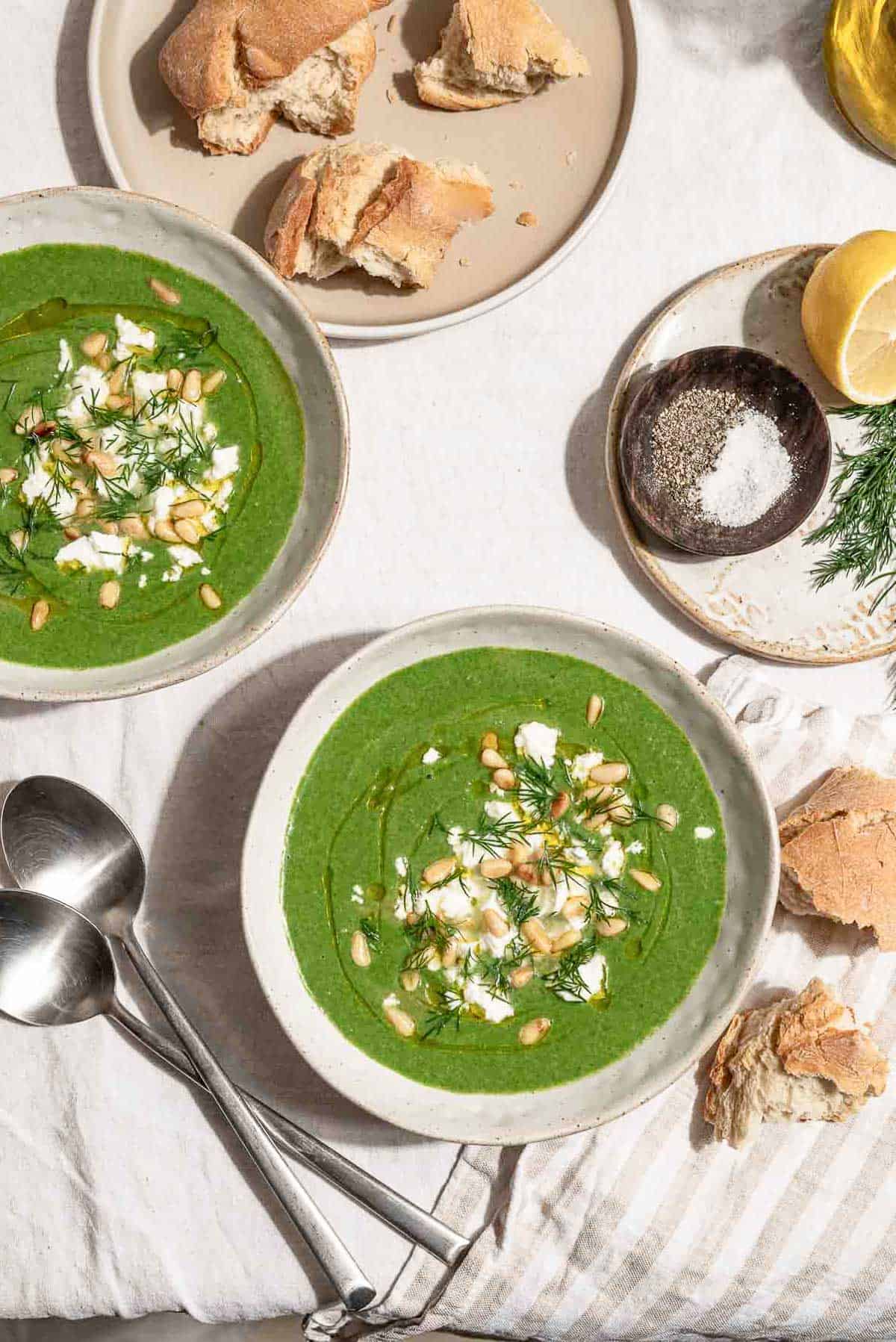 An overhead photo of 2 bowls of spinach soup topped with dill, pine nuts, crumbled feta and a drizzle of olive oil. Next to these is a cloth napkin, 2 spoons, pieces of crusty bread, and plate with a sprig of dill, a bowl of salt and pepper and a lemon half.