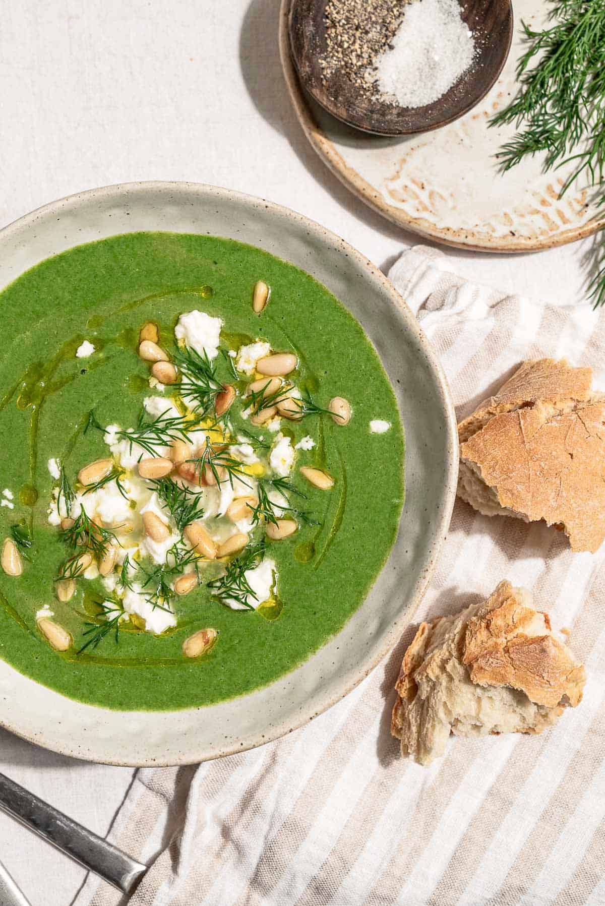 An overhead photo of a serving of spinach soup in a bowl topped with dill, pine nuts, crumbled feta and a drizzle of olive oil. Next to this is a cloth napkin, pieces of crusty bread, a sprig of dill, and a bowl of salt and pepper.