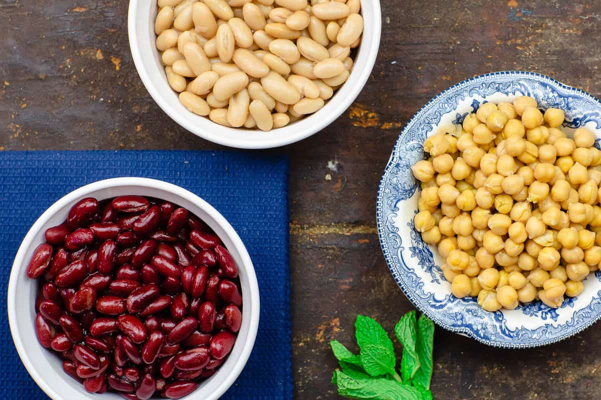 An overhead photo of a bowl of cannellini beans, a bowl of kidney beans, a bowl of chickpeas and a sprig of mint.