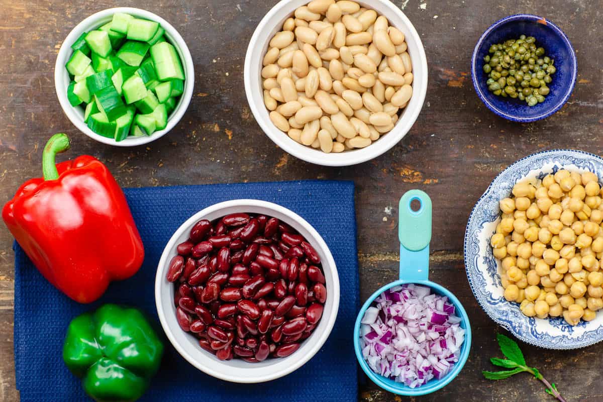 The ingredients for a Mediterranean three bean salad including kidney beans, chickpeas, green bell pepper, red bell pepper, diced red onion, and diced cucumber.