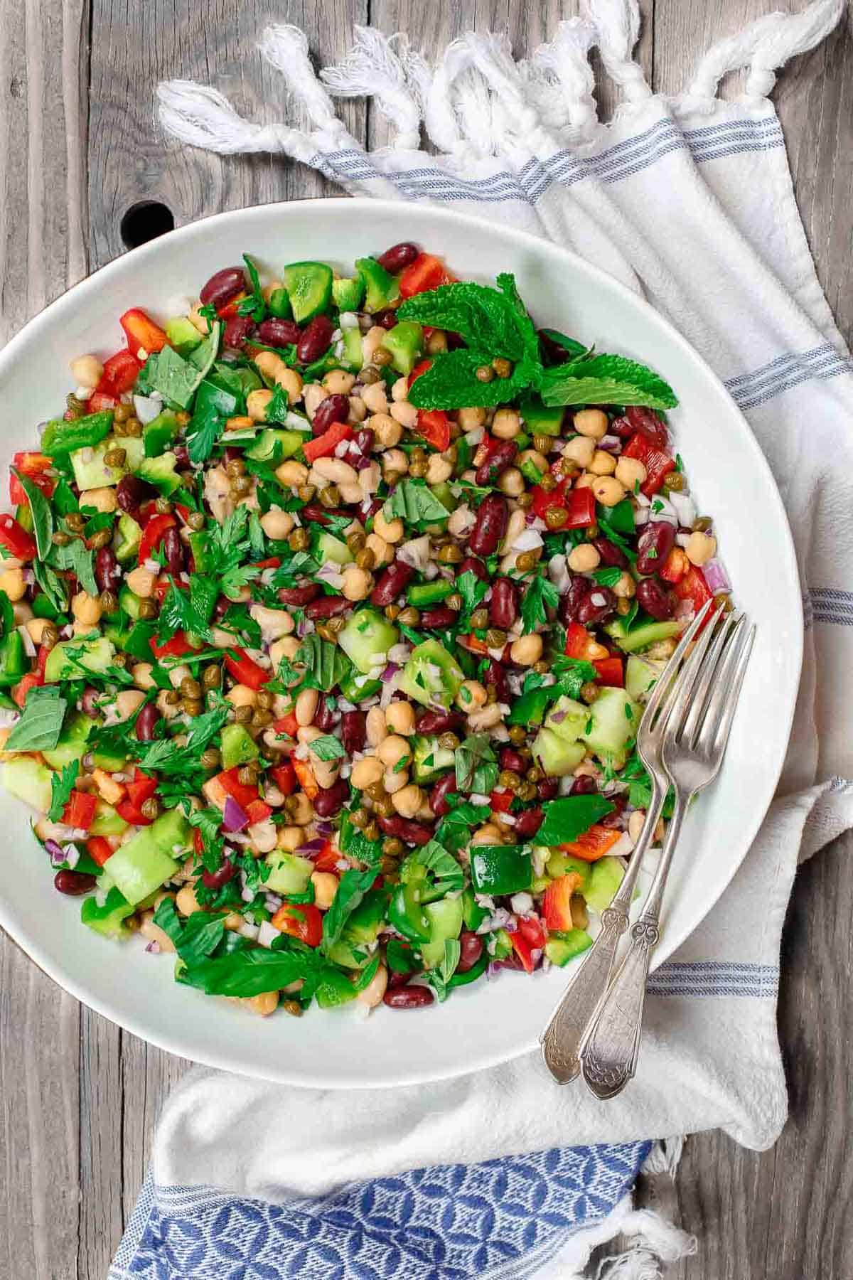 An overhead photo of a Mediterranean three bean salad in a serving bowl with two forks. Next to this is a kitchen towel.