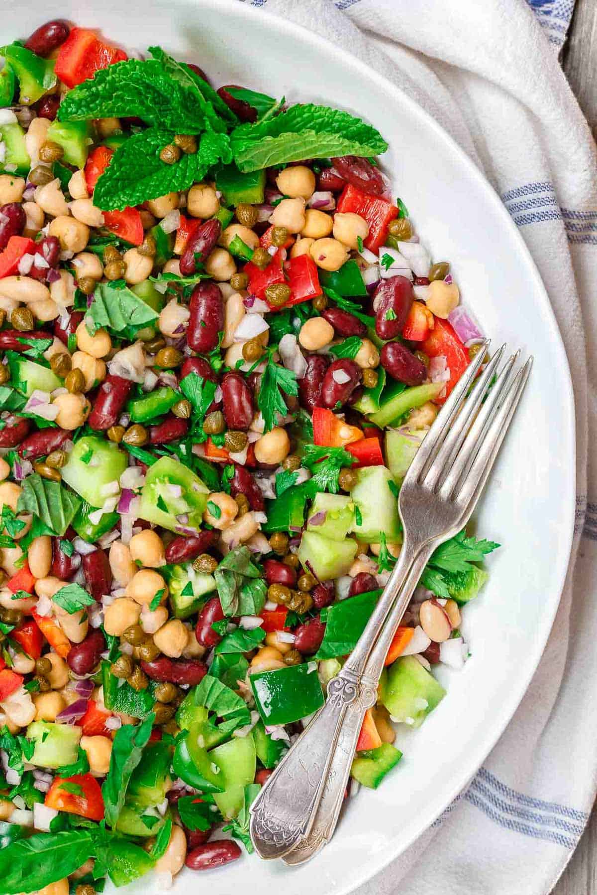 An overhead photo of a Mediterranean three bean salad in a serving bowl with two forks. Next to this is a kitchen towel.