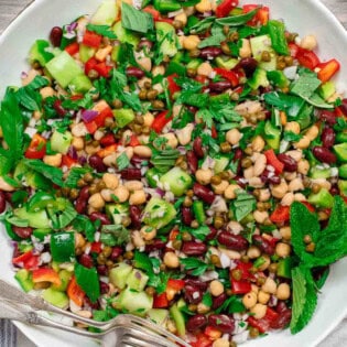 An overhead photo of a Mediterranean three bean salad in a serving bowl with two forks. Next to this is a kitchen towel.