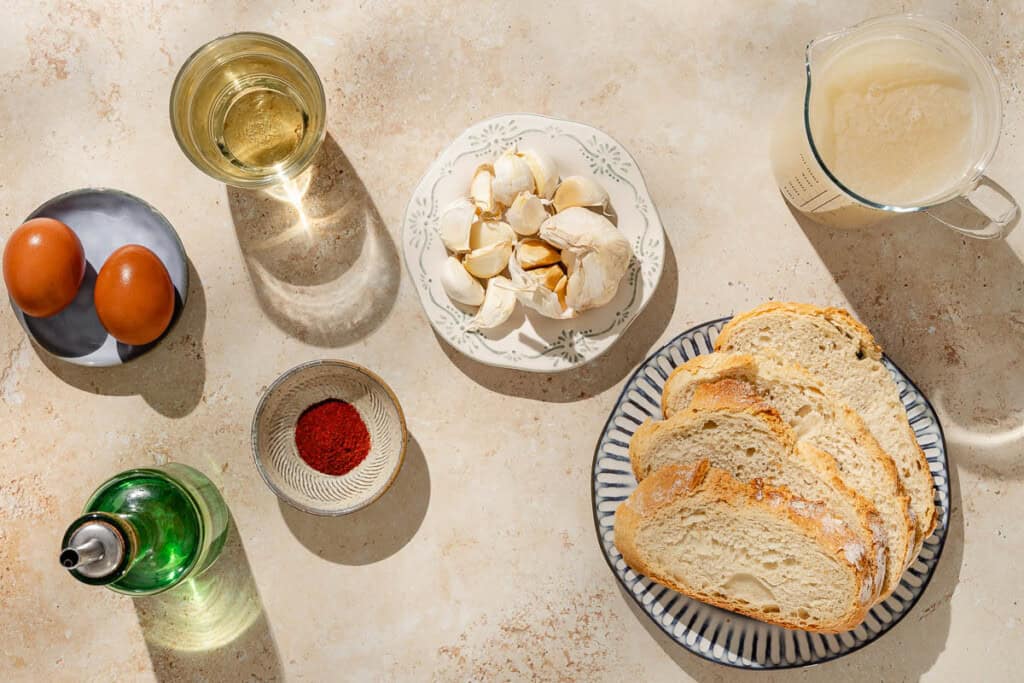 Ingredients for the sopa de ajo recipe including slices of bread, several garlic cloves, eggs, smoked paprika, dry white wine, and chicken stock.