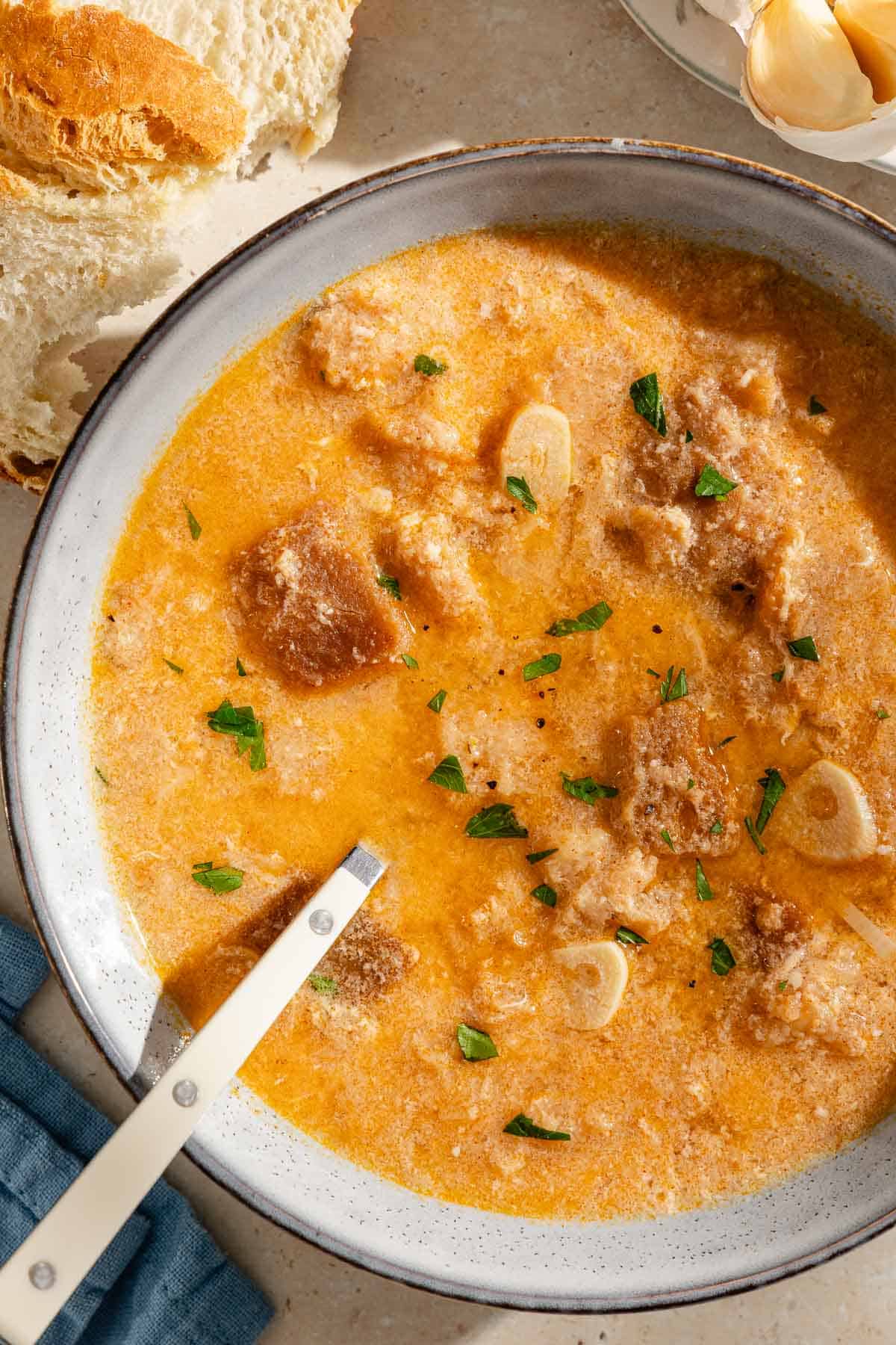 An overhead photo of a bowl of sopa de ajo with a spoon. Next to this is a cloth napkin, some crusty bread, and some garlic cloves.