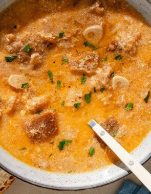 An overhead photo of a bowl of sopa de ajo with a spoon. Next to this is a cloth napkin, some crusty bread, a bowl of smoked paprika and a plate with garlic cloves.