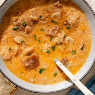 An overhead photo of a bowl of sopa de ajo with a spoon. Next to this is a cloth napkin, some crusty bread, a bowl of smoked paprika and a plate with garlic cloves.