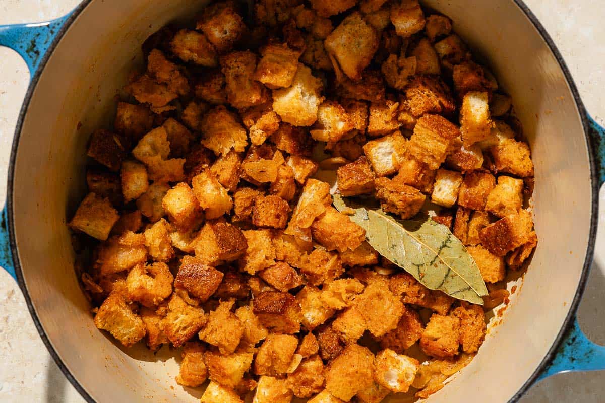 An overhead photo of the toasted bread cubes and a bay leaf after being added to the sauteed sliced garlic in a large pot.