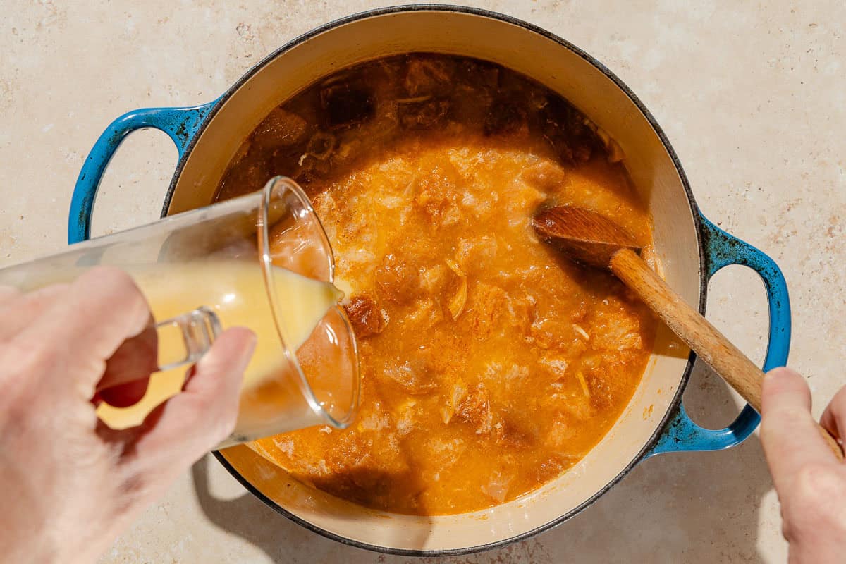 An overhead photo of the chicken stock being poured into the large pot with the other ingredient while being stirred in with a wooden spoon.