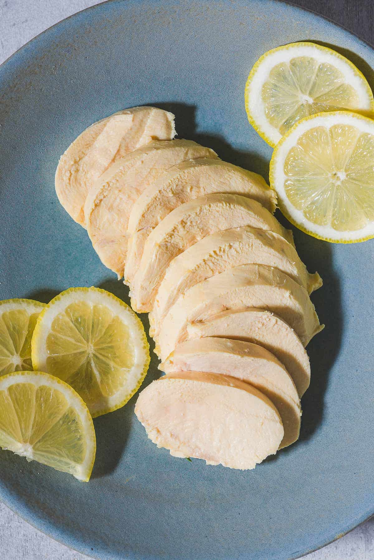 An overhead photo of a sliced poached chicken breast on a plate with lemon slices.